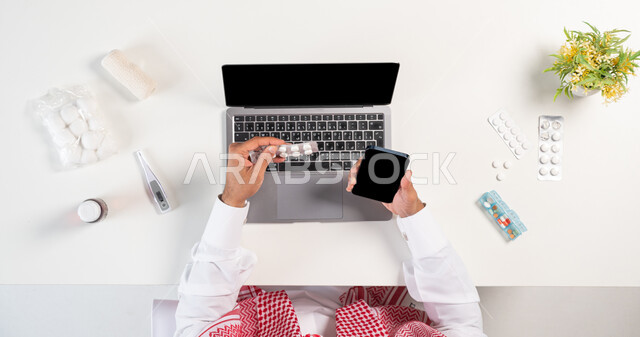 استخدام التقنيات الحديثة لطلب المستلزمات الدوائية، الحصول على مجموعة منوعة من الدواء، بورتريه علوي عامودي لرجل عربي خليجي سعودي يرتدي الثوب التقليدي والشماغ يستخدم الهاتف والحاسوب بجانبه مجموعة من الأدوية، مفهوم التسوق عبر المواقع الالكترونية، خلفية بيضاء