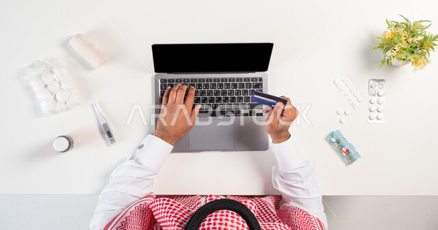 الحصول على مجموعة منوعة من الدواء عبر الإنترنت، الدفع من خلال بطاقة الصراف البنكية، بورتريه علوي عامودي لرجل عربي خليجي سعودي يرتدي الثوب التقليدي والشماغ يستخدم الحاسوب بجانبه مجموعة من الأدوية، استخدام التقنيات الحديثة لطلب المستلزمات الدوائية، خلفية بيضاء
