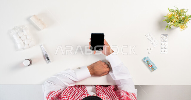 مفهوم التسوق عبر المواقع الالكترونية، بورتريه علوي عامودي لرجل عربي خليجي سعودي يرتدي الثوب التقليدي والشماغ يستخدم الهاتف المحمول وبجانبه مجموعة من الأدوية، استخدام التقنيات الحديثة لطلب المستلزمات الدوائية، خلفية بيضاء