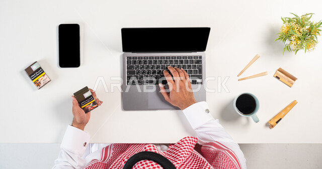 عادة ضارة تسبب الضرر بالصحة، تعاطي مواد سامة، أضرار التدخين على الجسد، شراء السجائر عبر الأونلاين، بورتريه علوي عامودي لرجل عربي خليجي سعودي يرتدي الثوب التقليدي والشماغ يستخدم الحاسب المحمول وبجانبه علب الدخان والتبغ، الإدمان على التبغ والسجائر، خلفية بيضاء