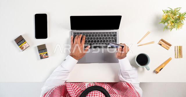 تعاطي مواد سامة، أضرار التدخين على الجسد، شراء السجائر عبر الأونلاين، بورتريه علوي عامودي لرجل عربي خليجي سعودي يرتدي الثوب التقليدي والشماغ يستخدم الحاسوب بجانبه علب الدخان والتبغ، الدفع من خلال بطاقة الصراف البنكية، عادة ضارة تسبب الضرر بالصحة، خلفية بيضاء