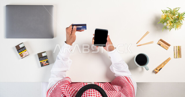 The harmful effects of smoking on the body, buying cigarettes online, a vertical portrait of a Saudi Gulf Arab man wearing a traditional dress and shemagh, using a phone with smoke and tobacco cans next to him, paying using a bank debit card, using toxic substances, a harmful habit that causes damage to health, white background.