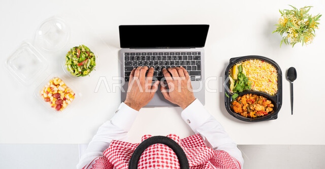 Using modern and advanced technologies, obtaining balanced nutrition through the Internet, a portrait of a Saudi Gulf Arab man using a laptop with plates of healthy food next to him, searching for an integrated health program through websites, maintaining physical health, white background