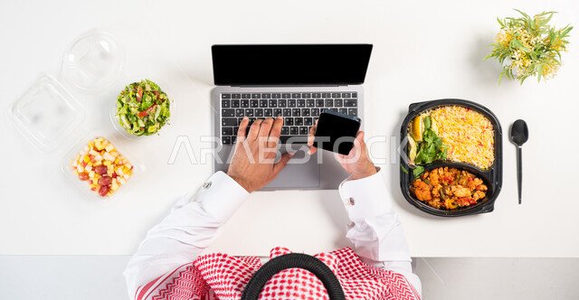 الحصول على الغذاء المتوازن عبر الأونلاين، بورتريه علوي عامودي لرجل عربي خليجي سعودي يرتدي الثوب التقليدي والشماغ يستخدم الهاتف والحاسوب وبجانبه أطباق من الطعام الصحي، استخدام التقنيات المتطورة، البحث عن برنامج صحي متكامل، الحفاظ على الصحة البدنية، خلفية بيضاء