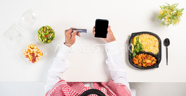 البحث عن برنامج صحي متكامل، الحفاظ على الصحة البدنية، بورتريه علوي عامودي لرجل عربي خليجي سعودي يرتدي الثوب التقليدي والشماغ يستخدم الهاتف وبجانبه أطباق من الطعام الصحي، الدفع من خلال بطاقة الصراف البنكية، الحصول على الغذاء المتوازن عبر الأونلاين، خلفية بيضاء