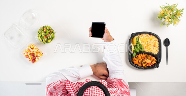 الحفاظ على الصحة البدنية، البحث عن برنامج صحي متكامل،  بورتريه علوي عامودي لرجل عربي خليجي سعودي يرتدي الثوب التقليدي والشماغ يستخدم الهاتف وبجانبه أطباق من الطعام الصحي، الحصول على الغذاء المتوازن عن طريق الأونلاين، عرض شاشة سوداء فارغة، خلفية بيضاء