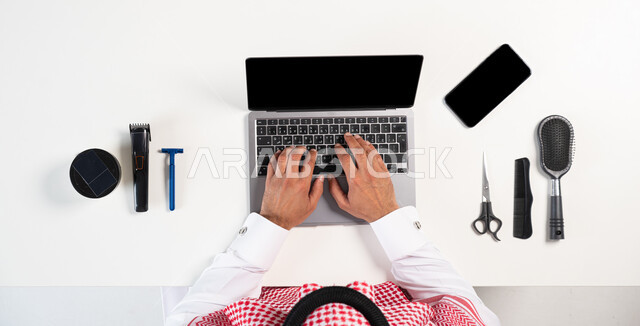 مفهوم التسوق الالكتروني، الاستفادة من التكنولوجيا لطلب المستلزمات اللازمة، الحصول على أدوات الحلاقة الرجالية، بورتريه علوي عامودي لرجل عربي خليجي سعودي يرتدي الثوب التقليدي والشماغ يستخدم الحاسوب المحمول بجانبه الهاتف، عرض شاشة سوداء فارغة، خلفية بيضاء