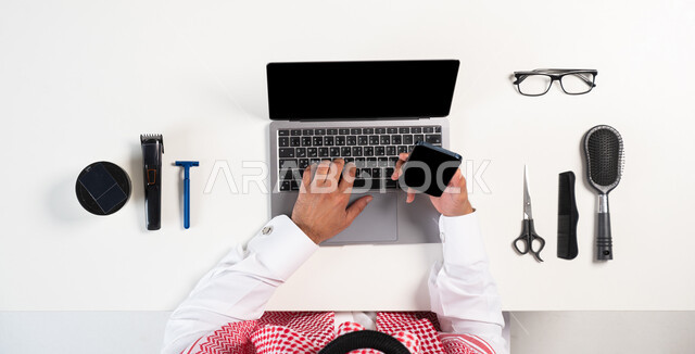 Taking advantage of technology to order the necessary supplies, obtain men’s shaving tools, a vertical overhead portrait of a Saudi Gulf Arab man wearing the traditional thobe and shemagh, using the phone and laptop, shopping through websites, displaying a blank black screen, white background