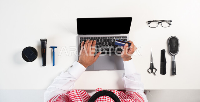 الحصول على أدوات الحلاقة الرجالية، بورتريه علوي عامودي لرجل عربي خليجي سعودي يرتدي الثوب التقليدي والشماغ يستخدم الحاسوب، الدفع عن طريق بطاقة الإئتمان البنكية، التسوق عبر المواقع الالكترونية، استخدام التقنيات الحديثة، عرض شاشة سوداء فارغة، خلفية بيضاء