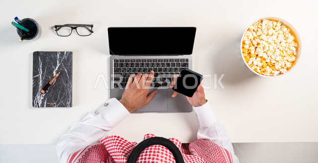The concept of entertainment with modern technology, a vertical overhead portrait of a Saudi Gulf Arab man wearing a traditional thobe and shemagh, using a computer and mobile phone with a book and medical glasses next to him, shopping through websites, buying and selling online, white background.