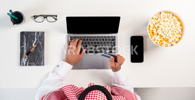 مفهوم التسوق  الإلكتروني، الحصول على المأكولات عبر المواقع الإلكترونية،  سهولة دفع الفواتير عبر بطاقة الصراف، بورتريه لرجل عربي خليجي سعودي يرتدي الثوب السعودي الخليجي يقوم باستخدام الحاسوب وبجانبه طبق من الطعام، خلفية بيضاء