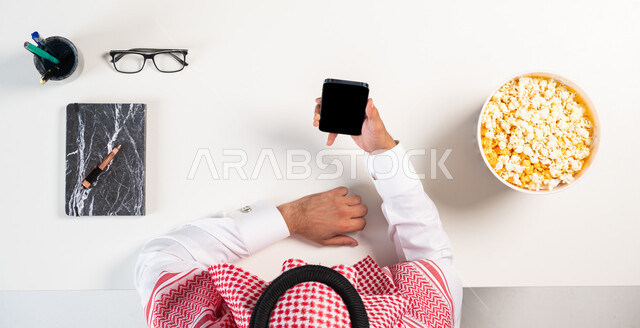 التسلية والترفيه مع الاستمتاع بطبق من الفوشار،  الحصول على المأكولات عبر المواقع الإلكترونية، بورتريه  علوي عامودي لرجل عربي خليجي سعودي يرتدي الثوب التقليدي يقوم باستخدام الهاتف المحمول، عرض شاشة سوداء فارغة، خلفية بيضاء