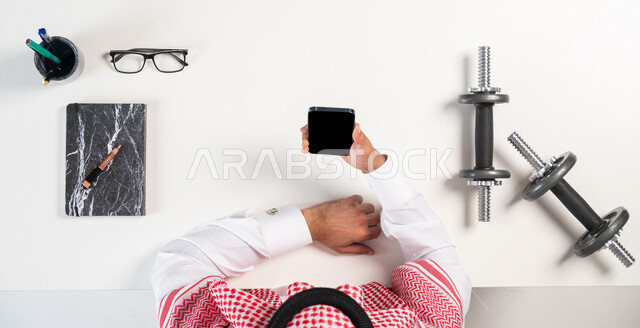 الحصول على المعدات الرياضية أونلاين، الاهتمام بالرشاقة واللياقة، بورتريه علوي عامودي لرجل عربي خليجي سعودي يرتدي الثوب التقليدي وبجانبه أثقال (الدمبلز)، عرض شاشة سوداء فارغة، خلفية بيضاء