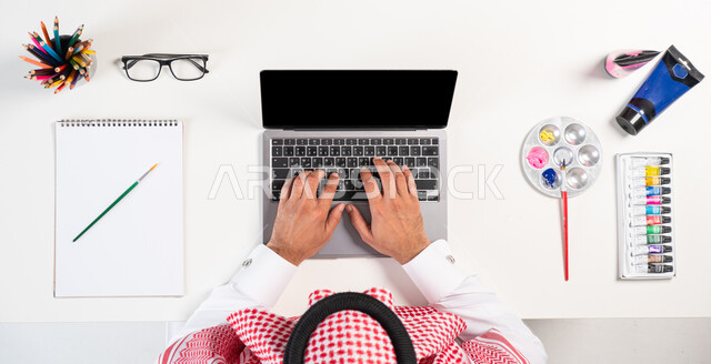 Using modern technologies, interest in activities and hobbies, practicing the talent of drawing and painting, a vertical overhead portrait of a Saudi Gulf Arab man wearing the traditional Saudi dress, using a computer, obtaining a drawing book and watercolors online, displaying a blank black screen, a white background.