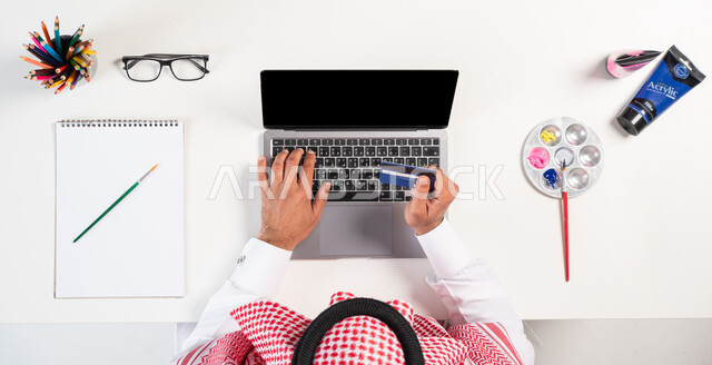 الاهتمام بالنشاطات والهوايات، ممارسة موهبة الرسم والتلوين، تسديد الفواتير عبر بطاقة الصراف الإلكترونية، بورتريه علوي عامودي لرجل عربي  خليجي سعودي يرتدي الثوب السعودي  التقليدي يقوم باستخدام الحاسوب، الحصول على دفتر الرسم والألوان المائية أونلاين، عرض شاشة سوداء فارغة، خلفية بيضاء