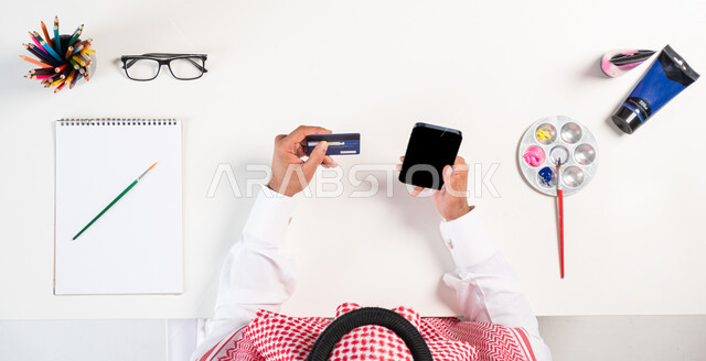 مفهوم التسوق الإلكتروني، سهولة دفع الفواتير  عبر البطاقة البنكية الإلكترونية، الاهتمام بالنشاطات والهوايات، ممارسة موهبة الرسم والتلوين، بورتريه علوي عامودي لرجل عربي  خليجي سعودي يرتدي الثوب التقليدي يقوم باستخدام الهاتف المحمول، الحصول على دفتر الرسم والألوان المائية أونلاين، عرض شاشة سوداء فارغة، خلفية بيضاء