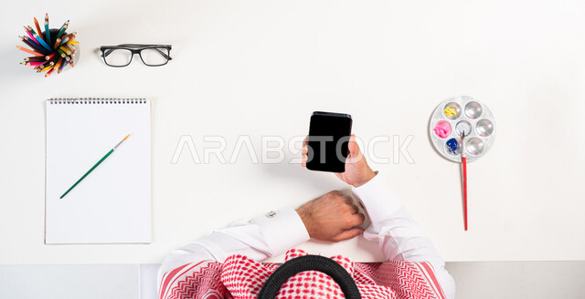 The concept of online shopping, interest in activities and hobbies, practicing the talent of drawing and painting, a vertical overhead portrait of a Saudi Gulf Arab man wearing a traditional dress, using a mobile phone, obtaining a drawing book and watercolors online, displaying a blank black screen, a white backgroundت