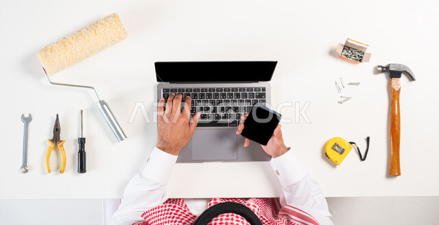 الاهتمام بالتجديد والتطور، الحصول على أدوات الصيانة والدهان عبر الإنترنت،سهولة شراء مستلزمات المهن أونلاين، بورتريه علوي عامودي لرجل عربي خليجي سعودي يرتدي الثوب التقليدي والشماغ يقوم باستخدام الهاتف المحمول والحاسوب، عرض شاشة سوداء فارغة، خلفية بيضاء