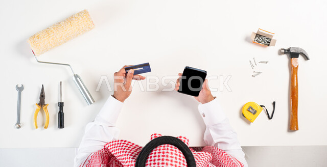 سهولة شراء مستلزمات المهن والدفع عبر البطاقة البنكية، الحصول على مجموعة من أدوات الصيانة والدهان عبر الإنترنت، طلب المهندسين والعمال من خلال المواقع الإلكترونية، بورتريه علوي عامودي لرجل عربي خليجي سعودي يرتدي الثوب التقليدي والشماغ يقوم باستخدام الهاتف المحمول وبجانبه معدات التصليح، الاهتمام بالتجديد والتطور، عرض شاشة سوداء فارغة، خلفية بيضاء