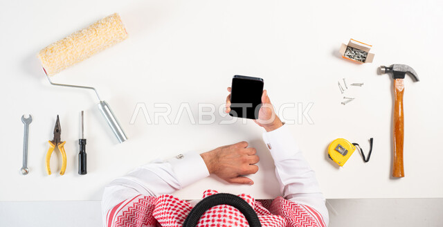 شراء أدوات الصيانة والدهان عبر الهاتف المحمول، بورتريه  علوي عامودي لرجل عربي خليجي سعودي يرتدي الثوب التقليدي والشماغ يستخدم الجوال وبجانبه معدات التصليح، الاهتمام بالتجديد والتطور، طلب العمال والمهندسين من خلال المواقع الإلكترونية، عرض شاشة سوداء فارغة، خلفية بيضاء