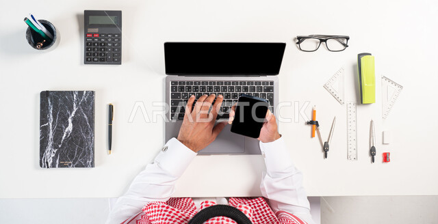 Obtaining school supplies online, purchasing mathematics tools through modern applications, a vertical overhead portrait of a Saudi Gulf Arab man wearing a traditional dress, using a mobile phone and computer,, displaying a blank black background, a white background