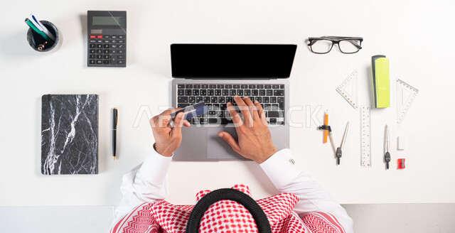 سهولة الشراء والدفع من خلال بطاقة الصراف البنكية، استخدام التقنيات الحديثة، التسوق الإلكتروني للحصول على أدوات الرياضيات أونلاين،  بورتريه  علوي عامودي لرجل عربي سعودي خليجي يرتدي الثوب التقليدي يقوم ياستخدام الحاسوب، عرض شاشة سوداء فارغة، خلفية بيضاء