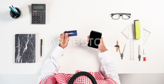 استخدام التقنيات الحديثة، الحصول على الأدوات الهندسية أونلاين،  بورتريه علوي عامودي لرجل عربي سعودي خليجي يرتدي الثوب لتقليدي يستخدم الهاتف المحمول، الدفع عبر بطاقة الصراف البنكية، الشراء عبر المواقع الإلكترونية، عرض شاشة سوداء فارغة، خلفية بيضاء