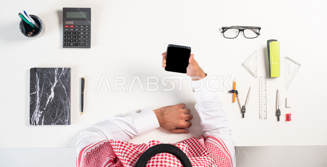 شراء أدوات الرياضيات والقرطاسية،  بورتريه علوي عامودي لرجل عربي خليجي سعودي يرتدي الثوب التقليدي يقوم باستخدام الهاتف المحمول، الحصول على المستلزمات المدرسية عبر الإنترنت، عرض شاشة سوداء فارغة، خلفية بيضاء