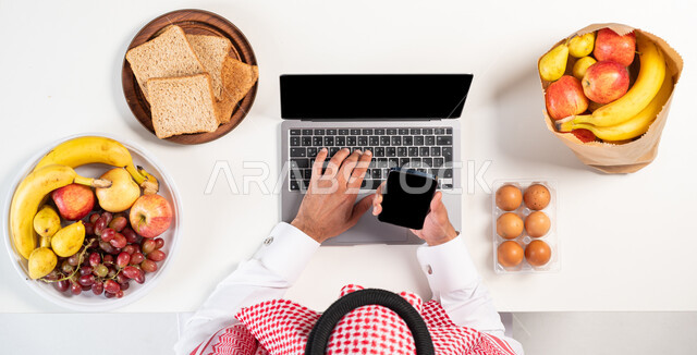 طلب المنتجات عبر التطبيقات الحديثة، بورتريه  علوي عامودي لرجل عربي خليجي سعودي يرتدي الثوب التقليدي يقوم باستخدام الهاتف المحمول والحاسوب، الحصول على أصناف من الفاكهة عبر الإنترنت، عرض خلفية سوداء فارغة، خلفية بيضاء