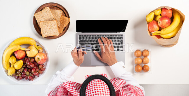 التسوق والشراء أونلاين، طلب المنتجات عبر المواقع الإلكترونية، دفع الفواتير عبر بطاقة الصراف البنكية، بورتريه علوي عامودي لرجل عربي خليجي سعودي يرتدي الثوب التقليدي يستخدم الحاسوب للحصول على أصناف من الفاكهة والتوست عبر الإنترنت، عرض خلفية شاشة سوداء فارغة، خلفية بيضاء