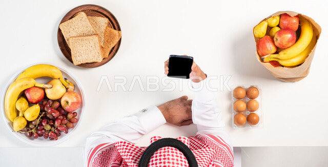 شراء الأغذية والأطعمة أونلاين،  بورتريه علوي عامودي لرجل عربي سعودي خليجي يرتدي الثوب التقليدي يستخدم الهاتف المحمول، استخدام أجهزة تقنية حديثة، الحصول على الغذاء الصحي والمتوازن، عرض شاشة سوداء فارغة، خلفية بيضاء