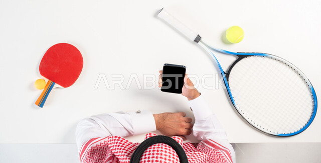 التسوق والشراء أونلاين، طلب المنتجات عبر التطبيقات الحديثة، بورتريه علوي عامودي لرجل عربي خليجي سعودي يرتدي الثوب التقليدي يقوم باستخدام جهاز الجوال وبجانبه مضارب التنس وكرة الطاولة، الحصول على الأدوات الرياضية والترفيهية عبر الإنترنت، عرض خلفية سوداء فارغة، خلفية بيضاء