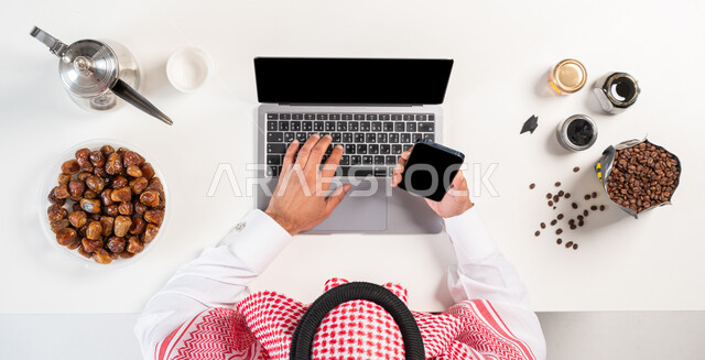 The concept of electronic shopping, obtaining food and drinks online, a vertical overhead portrait of a Saudi Gulf Arab man wearing a traditional dress, using a mobile phone and computer, using modern technical devices, purchasing through websites, displaying a blank black screen, a white background