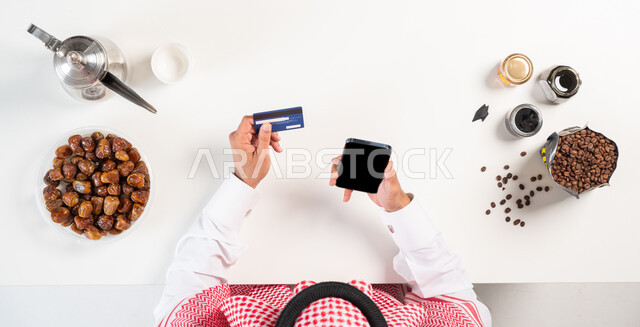 الحصول على المأكولات والمشروبات أونلاين، مفهوم التسوق الإلكتروني ،  بورتريه  علوي عامودي لرجل عربي سعودي خليجي يرتدي الثوب التقليدي يقوم باستخدام الهاتف المحمول وبجانبه دلة القهوة العربية مع طبق من التمر ، الدفع عبر بطاقة الصراف البنكية، بيع وشراء المنتجات الزراعية المحلية عبر الإنترنت، استخدام أجهزة تقنية حديثة، عرض شاشة سوداء فارغة، خلفية بيضاء