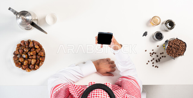 الضيافة التقليدية للمشروبات الساخنة، بورتريه علوي عامودي لرجل عربي سعودي خليجي يرتدي الثوب التقليدي يستخدم جهاز الهاتف المحمول وبجانبه دلة القهوة العربية مع طبق من التمر،بيع وشراء المنتجات الزراعية المحلية عبر الإنترنت، عرض شاشة سوداء فارغة، خلفية بيضاء