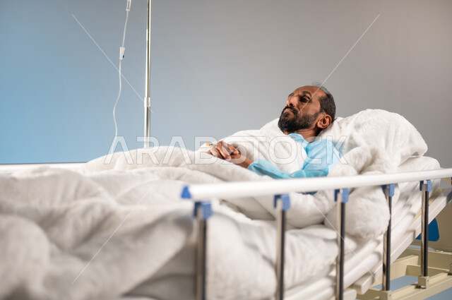 تلقي العلاج والتشخيص الطبي, اتباع الاجراءات الاحترازية من فايروس كورونا والحجر الصحي، مفهوم الطب والعناية الصحية، خدمات الرعاية الطبية في المستشفيات السعودية، بورتريه مقرب لرجل مريض عربي خليجي سعودي يرتدي يونيفورم العمليات مستلقي على السرير, خلفية زرقاء