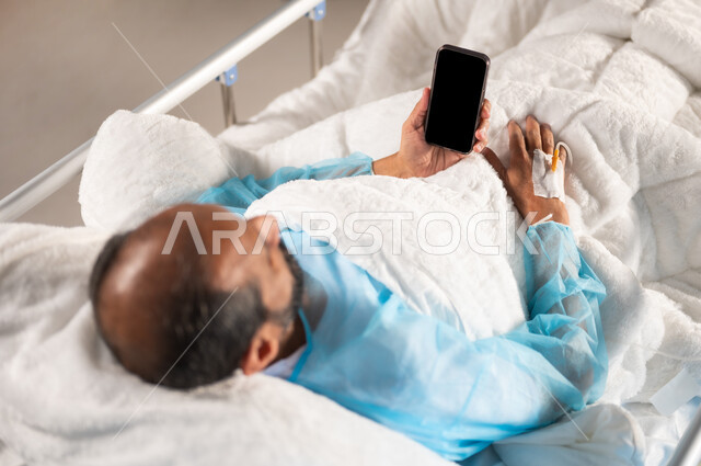 تبادل الأحاديث والأخبار مع الأهل والأصدقاء، إجراء مكالمة فيديو عبر الجوال، اتباع اجراءات الوقاية من فايروس كورونا والحجر الصحي, بورتريه علوي مقرب لرجل مريض عربي خليجي سعودي يرتدي يونيفورم العمليات مستلقي على السرير يقوم باستخدام الهاتف المحمول, خلفية زرقاء