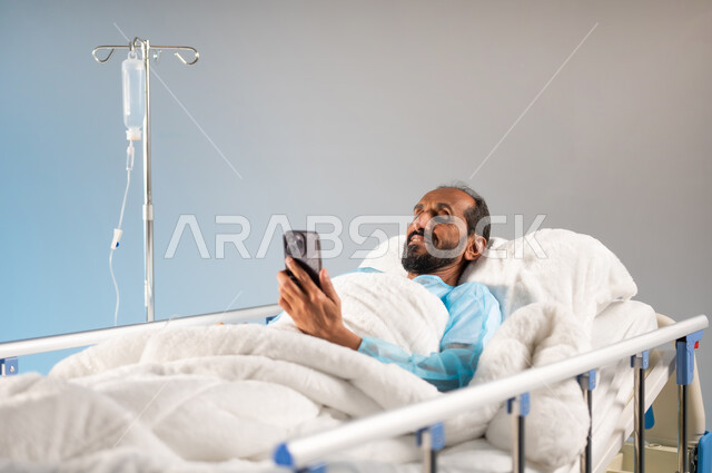 التسلية والترفيه عبر التطبيقات والبرامج الالكترونية، إجراء مكالمة فيديو عبر الجوال، اتباع اجراءات الوقاية من فايروس كورونا والحجر الصحي, بورتريه مقرب لرجل مريض عربي خليجي سعودي يرتدي يونيفورم العمليات مستلقي على السرير يقوم باستخدام الهاتف, خلفية زرقاء