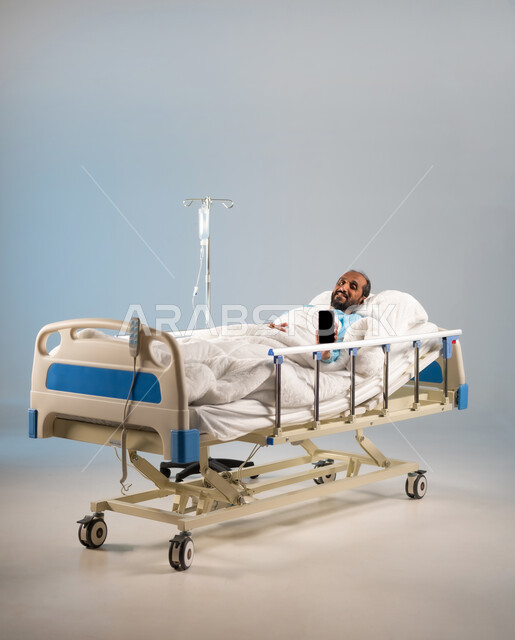 استخدام التكنولوجيا الحديثة والمتطورة، النظر الى الكاميرا بإيماءات الفرح والأمل، اتباع اجراءات الوقاية من فايروس كورونا والحجر الصحي, بورتريه لرجل مريض عربي خليجي سعودي يرتدي يونيفورم العمليات مستلقي على السرير يقوم باستخدام الهاتف, خلفية زرقاء