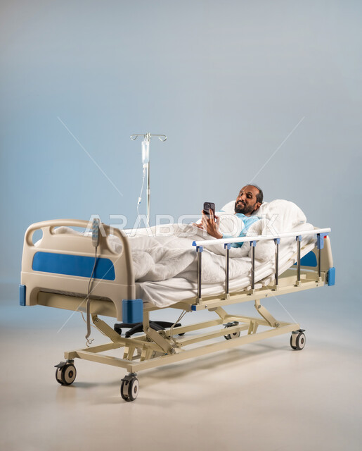 التقاط صور سيلفي تذكارية، إجراء مكالمة فيديو عبر الجوال، استخدام التكنولوجيا الحديثة والمتطورة، اتباع اجراءات الوقاية من فايروس كورونا والحجر الصحي, بورتريه لرجل مريض عربي خليجي سعودي يرتدي يونيفورم العمليات مستلقي على السرير يقوم باستخدام الهاتف, خلفية زرقاء