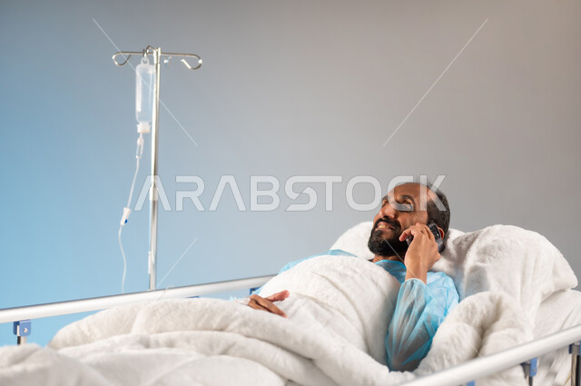 Using modern and advanced devices, making a phone call, following coronavirus prevention and quarantine measures, receiving treatment and health care, a close-up portrait of a sick Saudi Arabian Gulf man wearing an operating uniform lying on the bed using the phone, blue background
