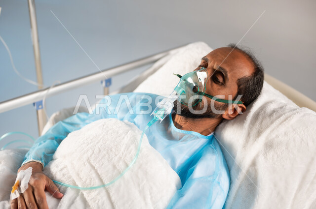 الحجر الصحي واتباع اجراءات الوقاية من فايروس كورونا، تلقي العلاج والتشخيص الطبي في المستشفى, بورتريه مقرب لرجل مريض عربي خليجي سعودي يرتدي يونيفورم العمليات مستلقي على السرير يضع كمامة الأكسجين, خدمات الرعاية الطبية في المستشفيات السعودية، خلفية زرقاء