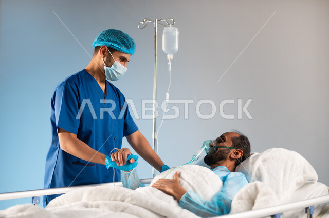 تبادل النظرات التي يملؤها التفاؤل والأمل والابتسامة، تلقي العلاج والرعاية الصحية، اتباع إجراءات الوقاية من فايروس كورونا، بورتريه مقرب لرجل عربي خليجي سعودي مستلقي على السرير يرتدي يونيفورم العمليات يضع كمامة الأكسجين و يمسك بيد الممرض, خلفية زرقاء
