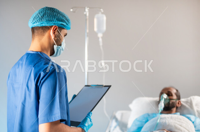 اتباع إجراءات الوقاية من فايروس كورونا، تقديم الاستشارات والخدمات العلاجية، إجراء الفحوصات والمتابعة الدورية, صورة ضبابية مقربة لممرض عربي خليجي سعودي يرتدي يونيفورم العمل والكمامة يقوم بتسجيل معلومات المريض, خدمات العناية الطبية في المستشفيات, خلفية زرقاء