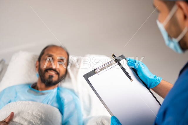 تقديم الاستشارات والخدمات العلاجية، إجراء الفحوصات والمتابعة الدورية للمرضى, صورة ضبابية مقربة لممرض عربي خليجي سعودي يرتدي يونيفورم العمل والكمامة يقوم بتسجيل وتدوين معلومات المريض, خدمات العناية الطبية في المستشفيات السعودية, خلفية زرقاء