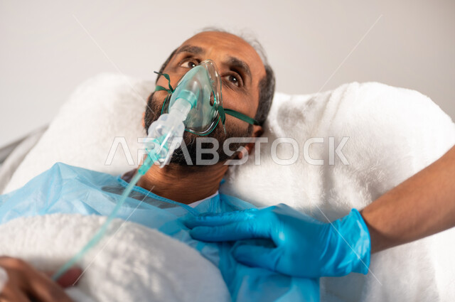 Looking and paying attention to the nurse to hear medical instructions, conducting periodic examinations and follow-ups for patients, medical care services in Saudi hospitals, a close-up portrait of a Saudi Gulf Arab man lying on the bed wearing an operating uniform and an oxygen mask, blue background