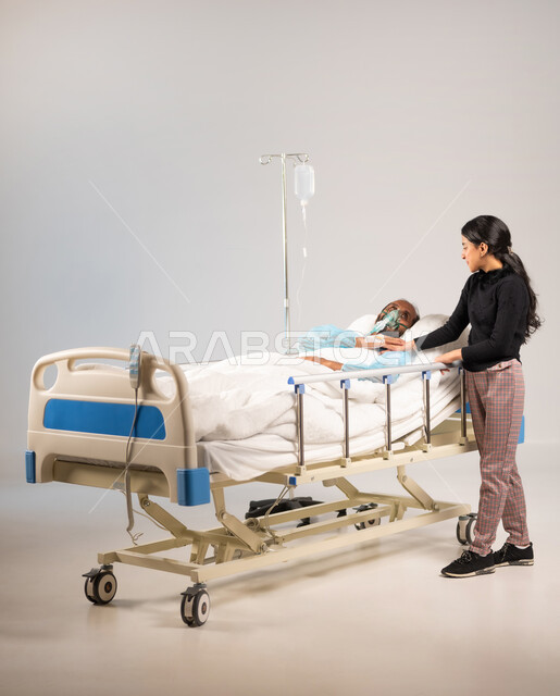 تبادل نظرات يملؤها الأمل والحب, المواساة والتخفيف عن المريض من خلال الامساك بيده، بورتريه لرجل عربي خليجي سعودي يرتدي يونيفورم العمليات مستلقي على السرير يضع كمامة الأكسجين ينظر الى ابنته, الرعاية الصحية والطب, تلقي العلاج والعناية في المستشفى, خلفية زرقاء