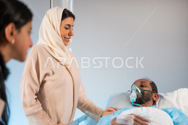 الاهتمام والإعتناء بالمريض ورعايته، شعور الشخص المصاب بالإرهاق والتعب وأهمية العناية به، بورتريه مقرب لرجل عربي خليجي سعودي يرتدي يونيفورم العمليات مستلقي على السرير يضع كمامة الأكسجين بجانبه زوجته وابنته مبتسمين, ايماءات العطف والحنان، خلفية زرقاء