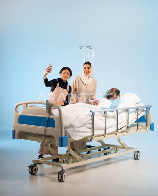 النظر الى الهاتف لالتقاط صور سيلفي تذكارية، استخدام الأجهزة والتقنيات الحديثة المتطورة، إجراء مكالمة فيديو عن طريق الجوال، بورتريه لرجل عربي خليجي سعودي يرتدي يونيفورم العمليات مستلقي على السرير يقفون بجانبه عائلته, مفهوم الطب والرعاية الصحية، خلفية زرقاء