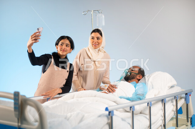 Using advanced modern devices and technologies, taking souvenir selfies via mobile phone, making a video call via mobile phone, a close-up portrait of a Saudi Gulf Arab man wearing an operating uniform lying on the bed with his family standing next to him, the concept of medicine and health care, blue background